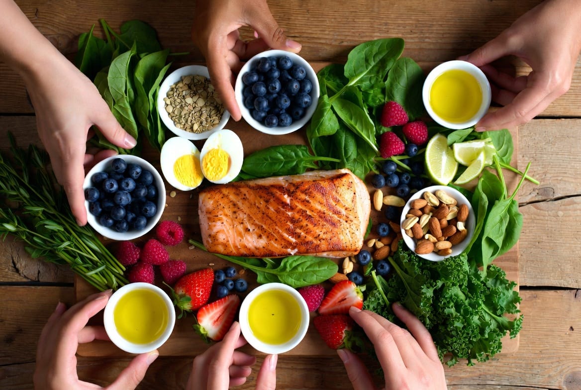 Supporting Image: Vibrant, appetizing flat-lay of recovery-boosting foods arranged artistically – grilled salmon fillet, boiled eggs, mixed nuts, fresh leafy greens, and colorful berries on a wooden surface with herbs and olive oil. Include diverse hands preparing or enjoying the meal to show community and action, complex composition with natural lighting and depth. Caption: "Practical, delicious foods that fuel brain healing and sustained recovery success."